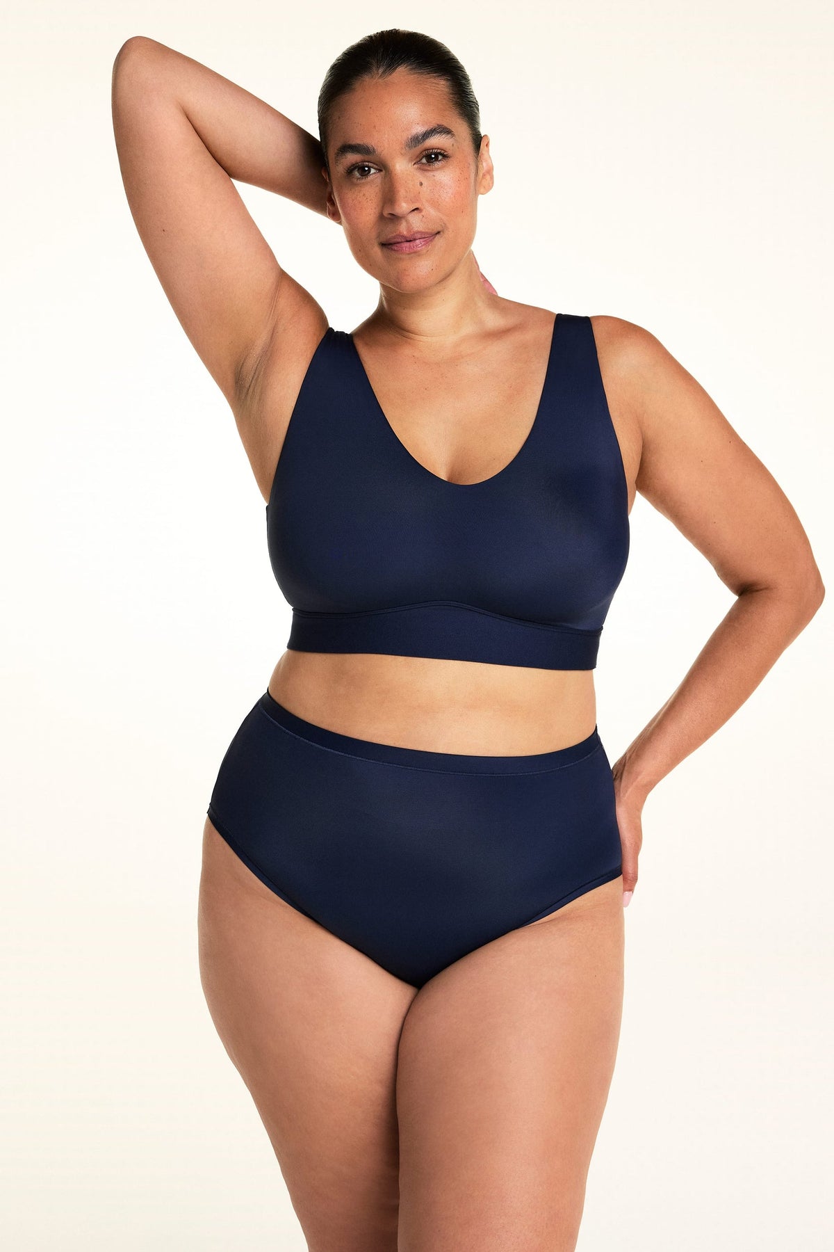 On a light studio backdrop, Jennifer wears the Bliss Highwaist in Navy with a matching navy bralette. Standing with one arm raised behind her head, she showcases the smooth high-rise waistband and full-coverage silhouette. sizing: Laura wears size Large