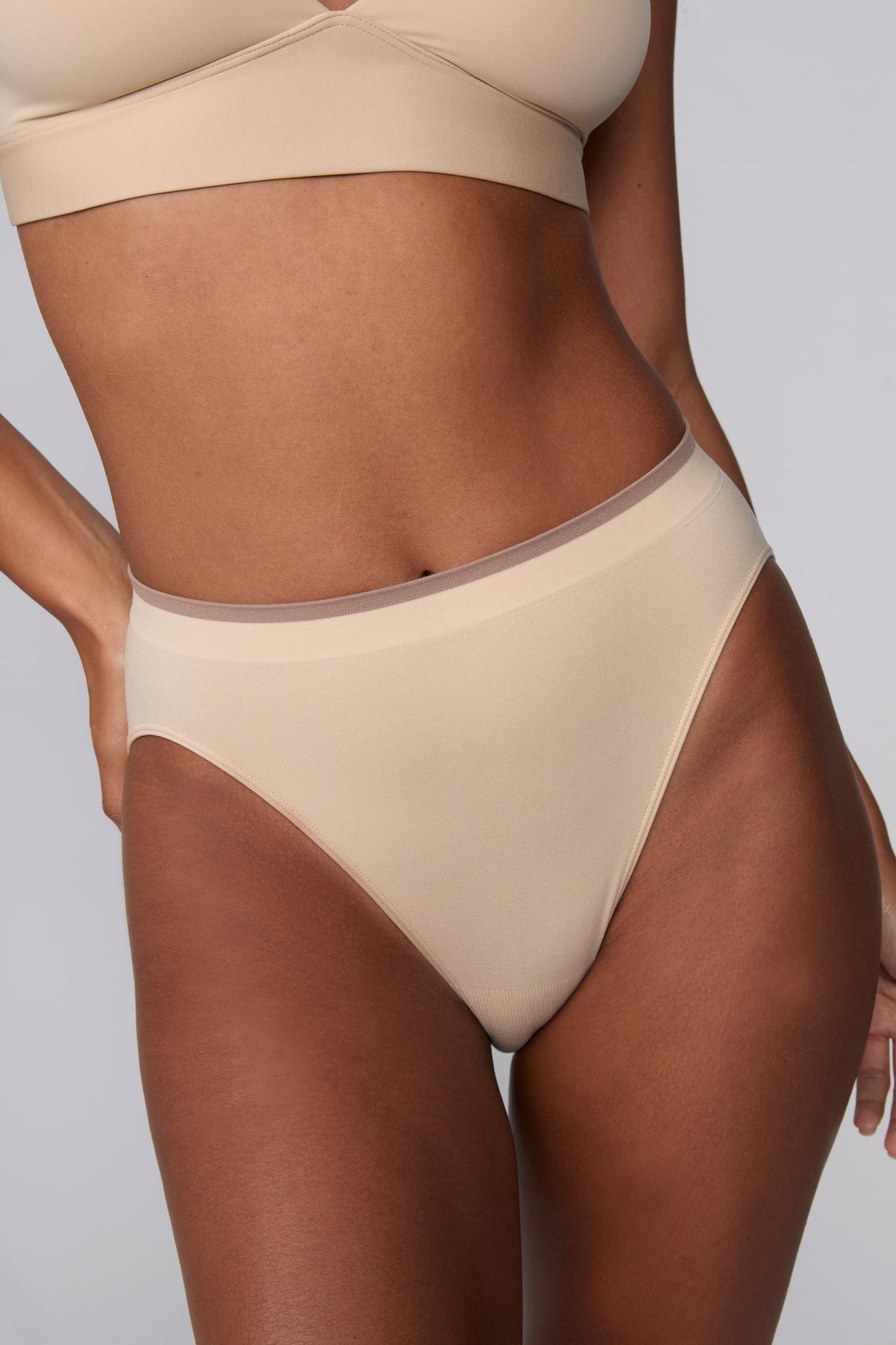In a studio setting, Courtney wears the Smooth Seamless Brief in Beige. A close-up front view highlights the smooth fabric, high-leg silhouette, and contrast trim waistband.