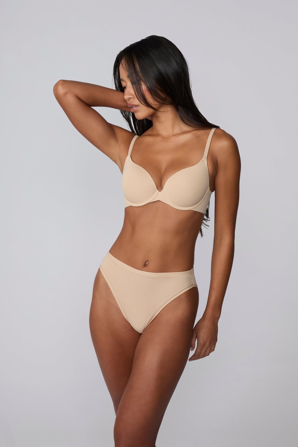 On a light grey studio background, Amber wears the Base Push-Up Bra in Beige with one arm raised. The molded, lightly lined push-up cups create lift and definition, finished with thin adjustable straps and a smooth microfiber band.
