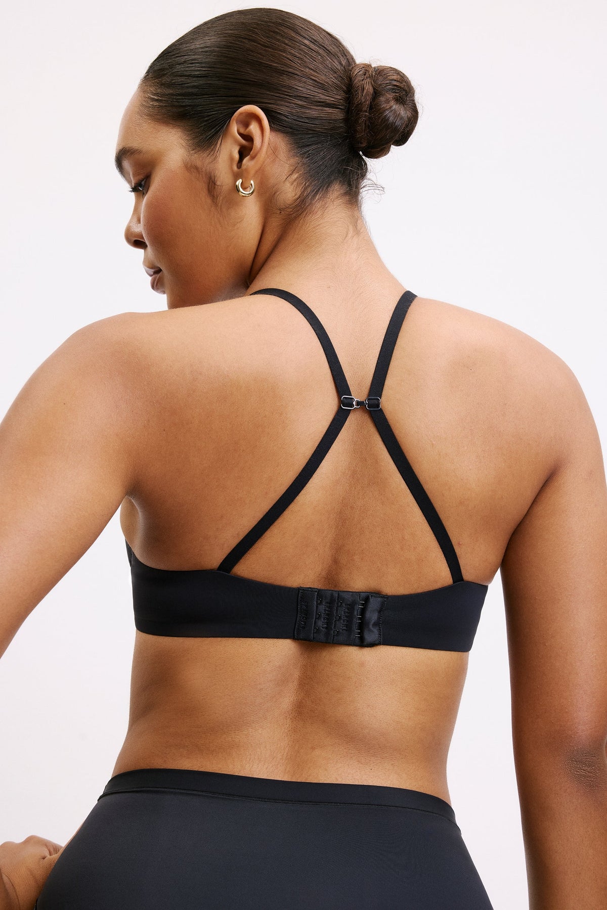 On a light studio backdrop, Amber wears the Base T-Shirt Bra in Black with the J-hook clasped, converting the adjustable straps into a racerback. The smooth microfiber band and hook-and-eye closure sit flat across her back.