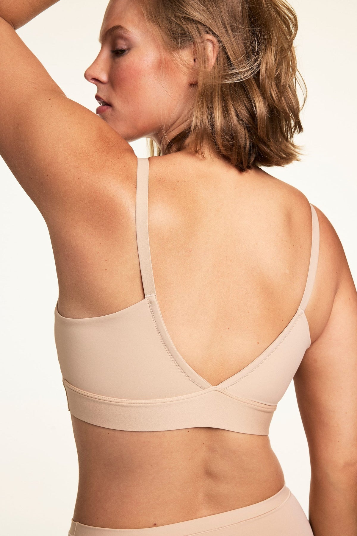 On a white studio backdrop, Sonnie wears the Bliss Triangle Bralette in Beige, shown from the back to highlight the adjustable straps, smooth back band, and wire-free design.