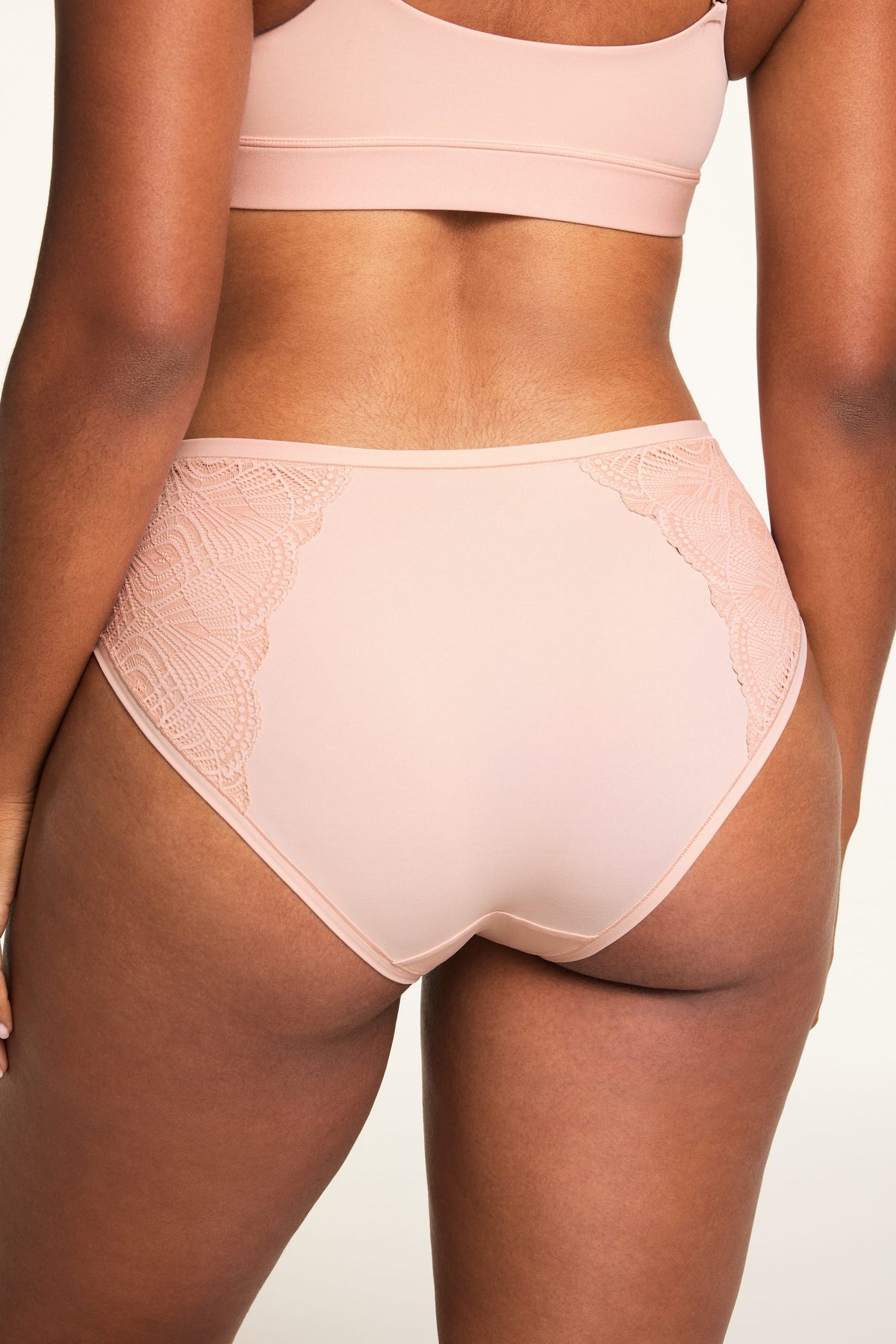 On a light studio backdrop, Amber wears the Bliss Lace Bralette and Bliss Lace Highwaist Brief in Ember Rose, shown from the back with the bralette’s thin straps and lace-trimmed details visible.