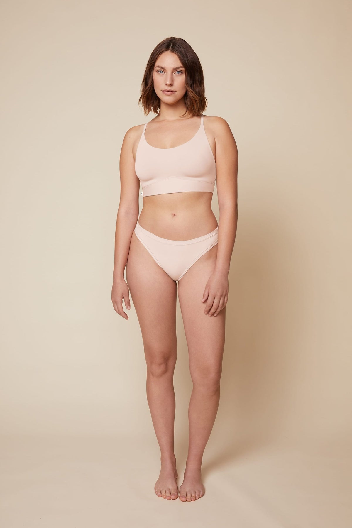 On a neutral beige backdrop, Carli wears the Bliss Racerback Bralette in Rose with coordinating underwear, showcasing the scoop neckline, adjustable straps, and supportive underband in a full-body front view. 