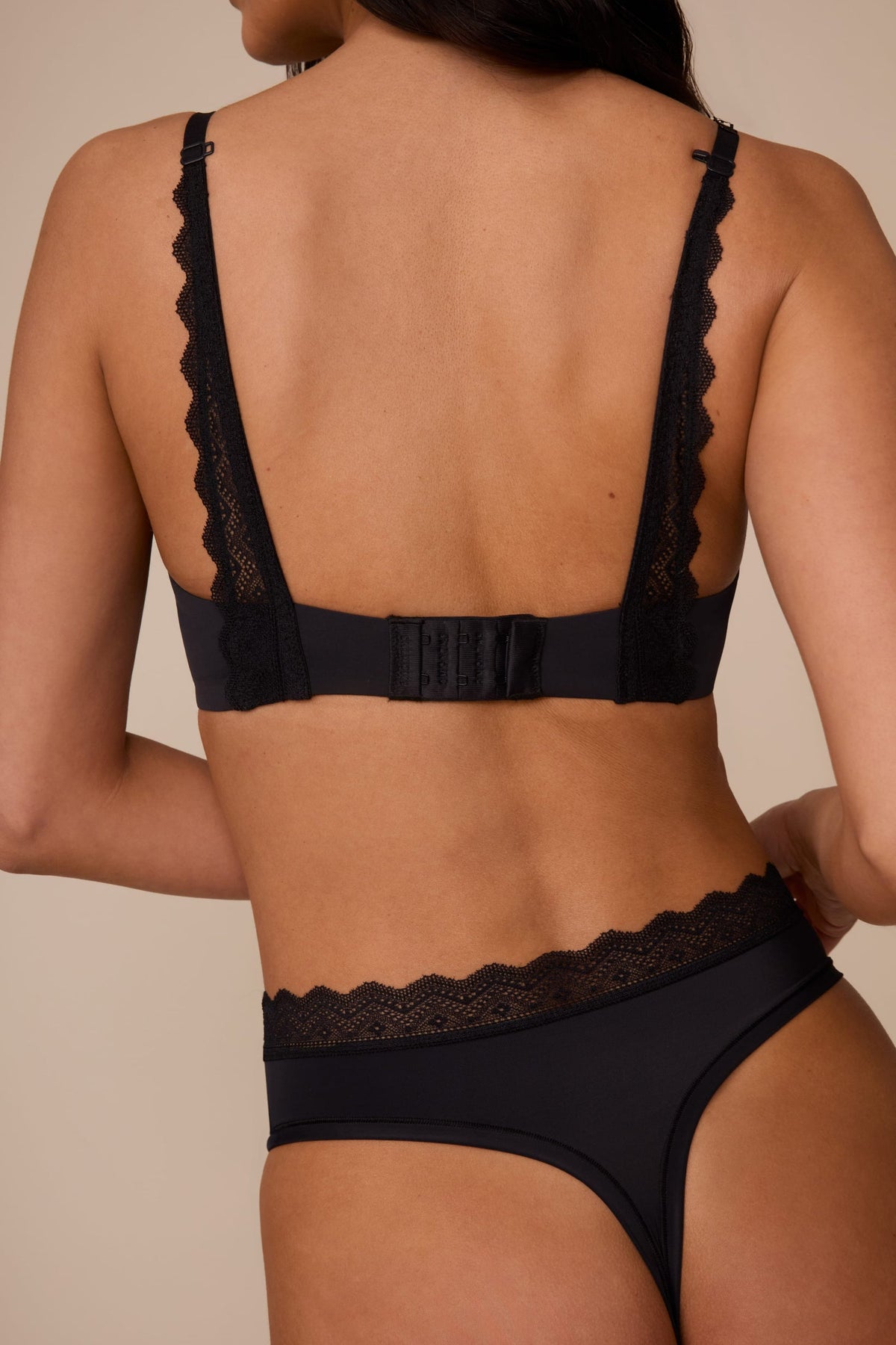 On a neutral studio backdrop, Marcela wears the Base Lace T-Shirt Bra in Black with matching underwear. The back view showcases the smooth band, adjustable straps, hook-and-eye closure, and lace detailing extending from the straps to the band.