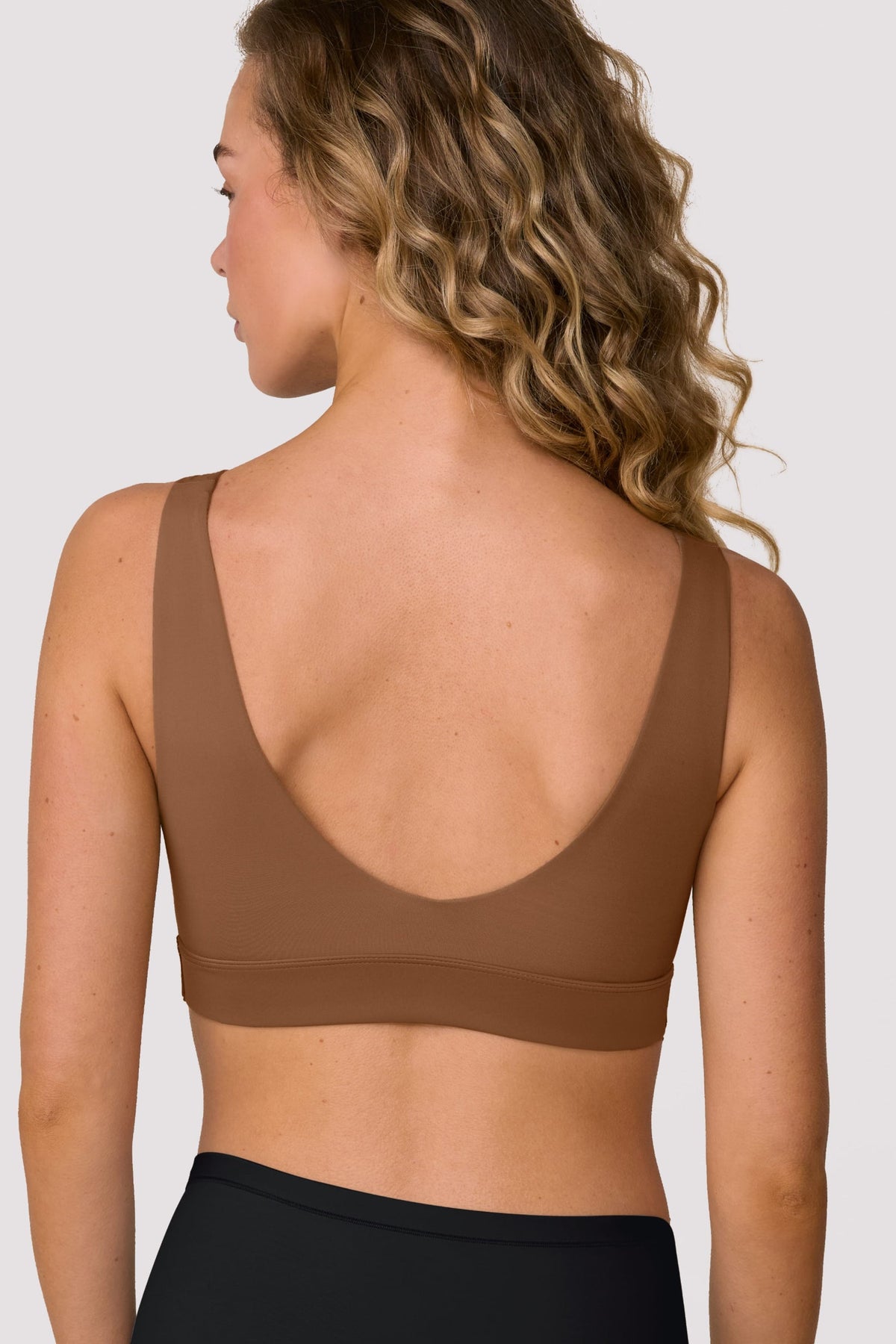 On a light studio backdrop, Freya wears the Bliss Bralette in Brown, shown from the back. The bralette features a scoop back, wide straps, and a smooth underband.