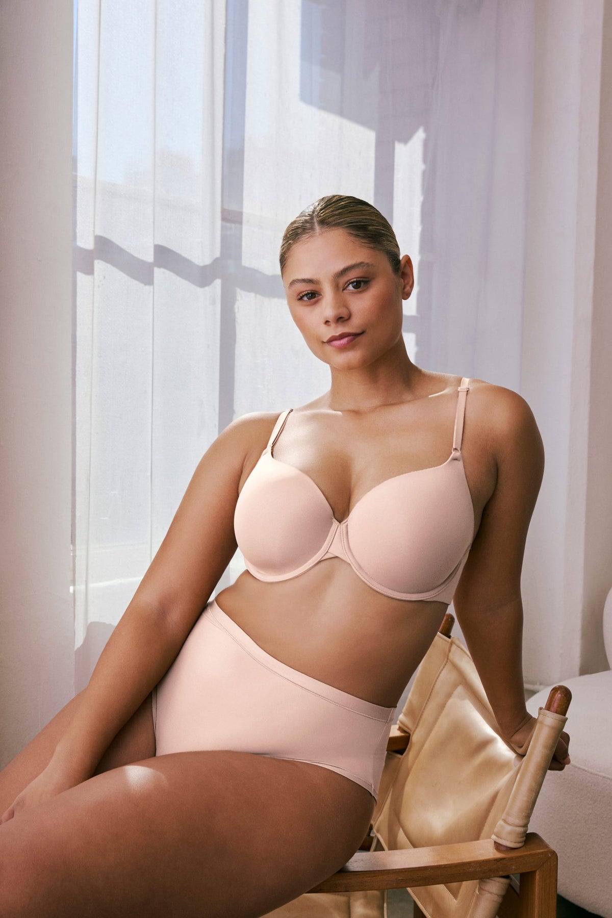 In a sunlit room, India wears the Base T-Shirt Bra in Rose with matching bottoms, seated on a chair. The bra features smooth molded cups, adjustable straps, and an underwire for support. sizing: India wears size 36D