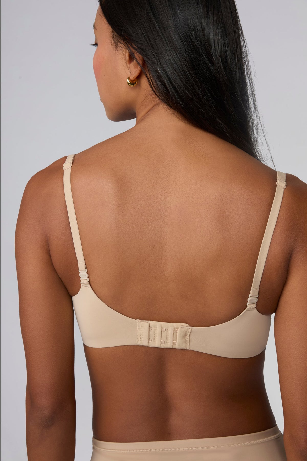 Against a light grey background, Amber models the Base Push-Up Bra in Beige from the back with straps worn straight. The adjustable straps and hook-and-eye closure are visible, with the soft microfiber band laying smooth across the back.