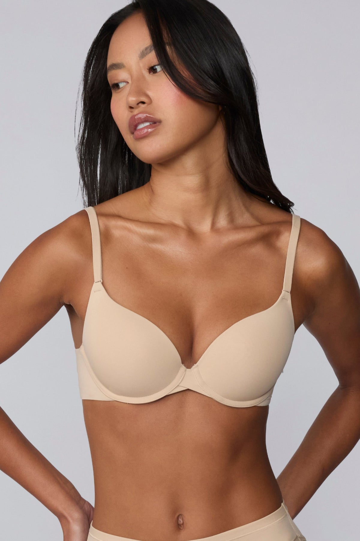 Against a light grey background, Amber models the Base Push-Up Bra in Beige, hands at her hips. The molded, lightly lined cups enhance lift and shape, with thin adjustable straps and smooth microfiber fabric for everyday comfort.