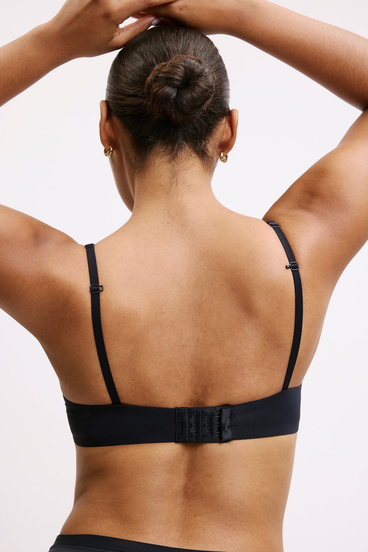 On a light studio backdrop, Amber faces away wearing the Base T-Shirt Bra in Black. The smooth microfiber band sits flat across her back, with adjustable straps and a hook-and-eye closure visible. Her hair is styled in a bun, highlighting the bra’s clean, supportive back design.