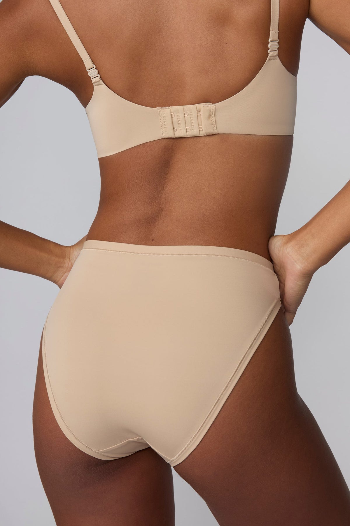 On a light gray studio backdrop, a back view of Amber wearing the Bliss French Cut Brief in Beige shows the smooth rear coverage and seamless-looking edges under the fabric.